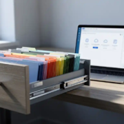 Geopende archieflade met kleurgecodeerde mappen naast laptop met cloud-interface op licht eiken bureau.