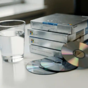 Halfvol glas water naast gestapelde, ongeopende Microsoft-softwaredozen op wit bureau, met cd's die uit een doos vallen.