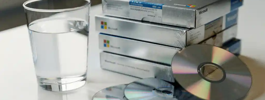Halfvol glas water naast gestapelde, ongeopende Microsoft-softwaredozen op wit bureau, met cd's die uit een doos vallen.