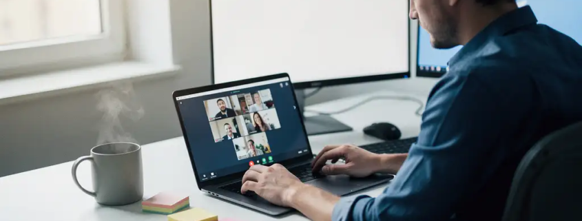 Professional aan modern bureau gebruikt Microsoft Teams op laptop, met tweede monitor, koffie en sticky notes in natuurlijk daglicht.