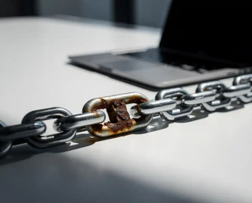 Roestige schakel in een stalen ketting op een kantoorbureau naast een laptop, symbool voor kwetsbaarheid in bedrijfsbeveiliging.