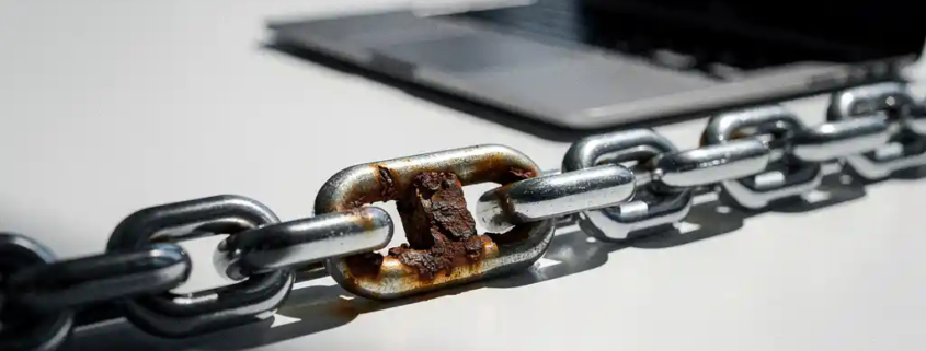 Roestige schakel in een stalen ketting op een kantoorbureau naast een laptop, symbool voor kwetsbaarheid in bedrijfsbeveiliging.
