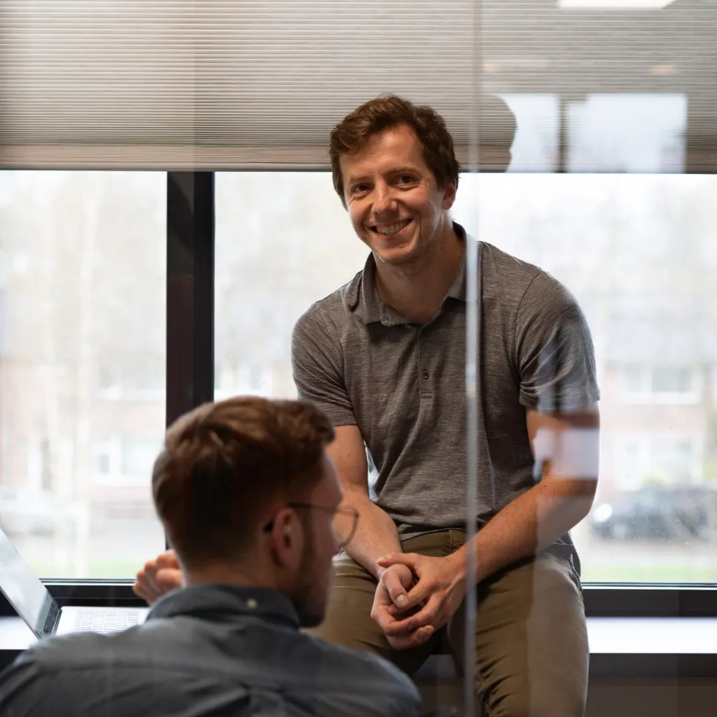 Twee medewerkers van Puur ICT zijn met elkaar in gesprek, waarbij één van hen op een bureau zit.
