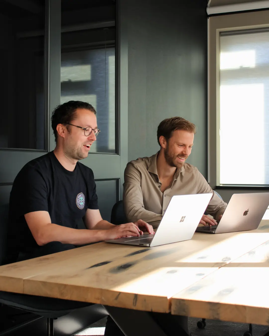 Twee medewerkers van Puur ICT zitten in een vergaderruimte en werken ieder op hun eigen laptop.