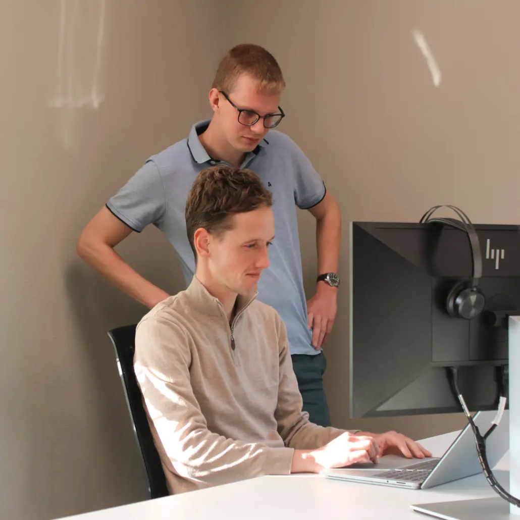 Twee medewerkers van Puur ICT kijken samen naar een computerscherm, één zittend en één staand achter het bureau.