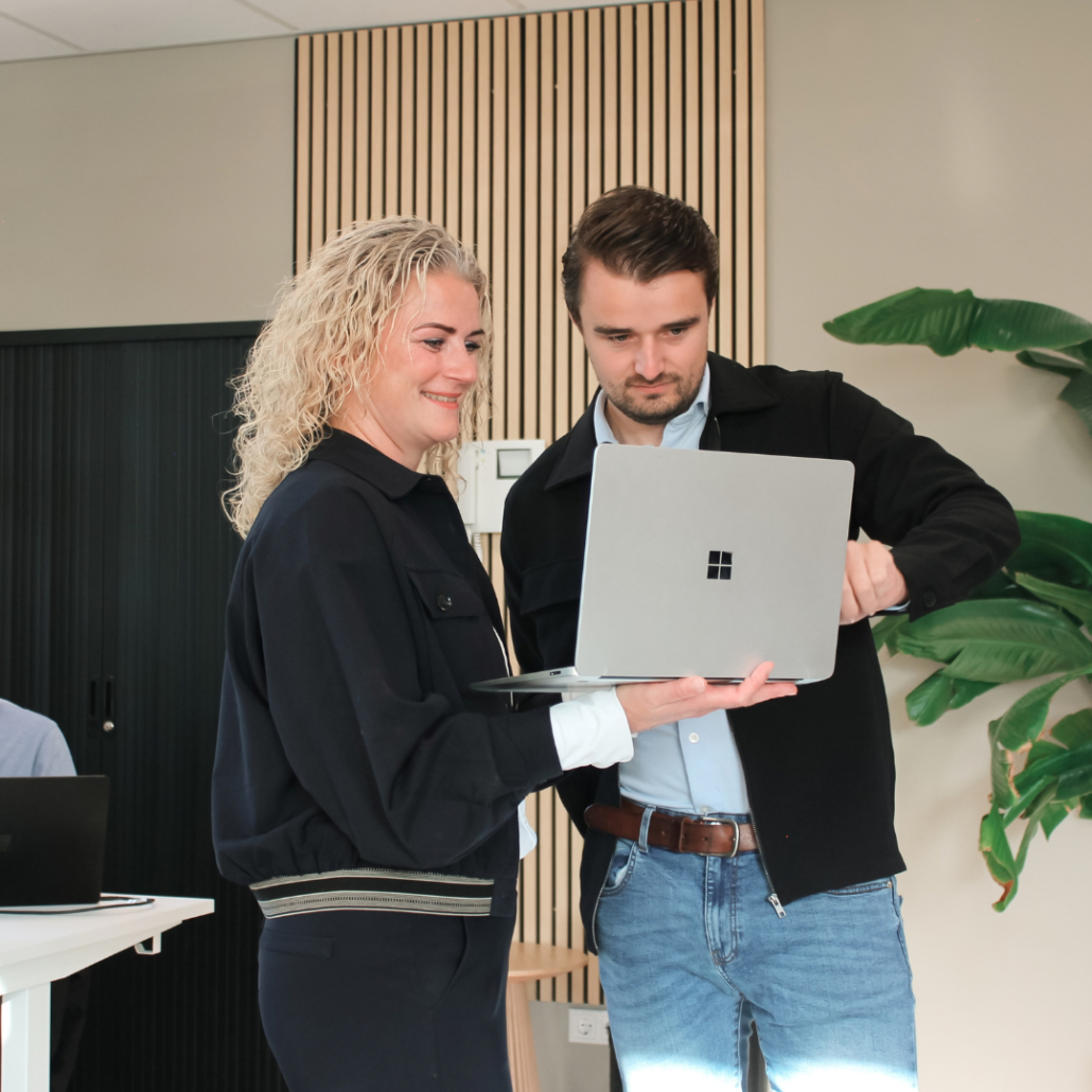 Mirjam & Wessel Twee collega's staan samen op kantoor en kijken aandachtig naar een laptopscherm. Ze overleggen terwijl ze door informatie op het scherm gaan in een moderne werkomgeving.