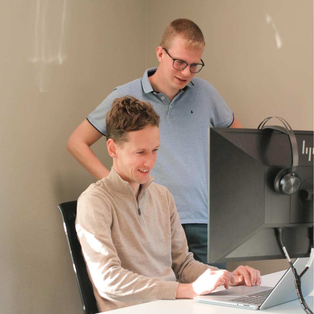Patrick & Jorrit Een collega staat achter een zittende collega en kijkt mee naar het scherm van diens laptop. Samen bespreken ze informatie op het scherm in een kantooromgeving.